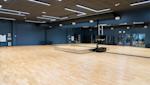 Rymligt träningsstudio med ljust trägolv, blå väggar, stora speglar och moderna LED-lampor i taket.