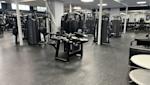 Modernt gym med svarta träningsapparater, vita pelare och grått golv under takbelysning.