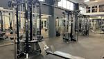 Modernt gym med svarta träningsstationer och kabelmaskiner, grått golv och stora fönster som släpper in naturligt ljus.