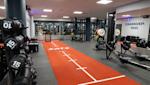 Modernt gym med orange löpbana, hantlar i förgrunden och träningsutrustning i bakgrunden under vitt tak.