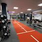 Modernt gym med orange löpbana, hantlar i förgrunden och träningsutrustning i bakgrunden under vitt tak.