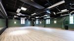 Hot studio med lyst tregulv, grønne vegger, svart tak med synlige rør og vinduer som slipper inn naturlig lys.
