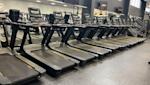 Rad av svarta löpband med digitala skärmar i ett modernt gym med stora fönster och TV-skärmar på väggen.
