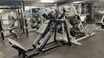Gym med benpress och viktmaskiner i svart och silver, lysrör i taket, grått golv och speglar på väggarna.