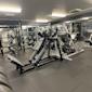 Gym med benpress och viktmaskiner i svart och silver, lysrör i taket, grått golv och speglar på väggarna.