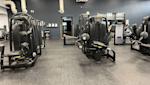 Modernt gym med svarta träningsapparater mot mörkgrå vägg, grå heltäckningsmatta och vit takpanel.