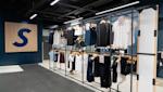 Modern klädbutik med svart industriellt tak, blå vägg med stor S-logotyp och klädställningar med t-shirts och byxor.