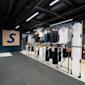 Modern klädbutik med svart industriellt tak, blå vägg med stor S-logotyp och klädställningar med t-shirts och byxor.
