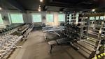 Modernt gym med hantlar, träningsbänkar och vikter. Svart tak, grått golv och stora fönster med grönska utanför.