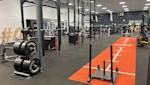 Modernt gym med power racks, viktskivor, orange träningsmatta och träningsutrustning i ett välupplyst lokalt.