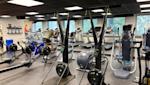 Modernt gym med roddmaskiner i förgrunden, löpband och styrketräningsutrustning, stora fönster och svart tak.