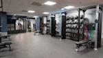 Modernt gym med träningsutrustning, hyllor med bollar och vikter, grått golv och vit takpanel med belysning.