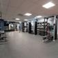 Modernt gym med träningsutrustning, hyllor med bollar och vikter, grått golv och vit takpanel med belysning.