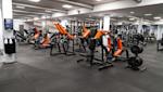 Modernt gym med orange och svarta träningsredskap, exponerade ventilationskanaler i taket och grått golvmatta.