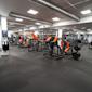 Modernt gym med orange och svarta träningsredskap, exponerade ventilationskanaler i taket och grått golvmatta.