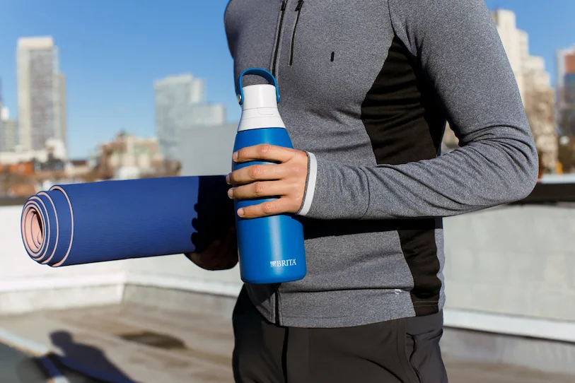 Person in athletic apparel, standing, holding rolled up yoga mat in one arm and Brita water bottle in other hand