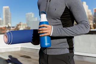 Person in athletic apparel, standing, holding rolled up yoga mat in one arm and Brita water bottle in other hand