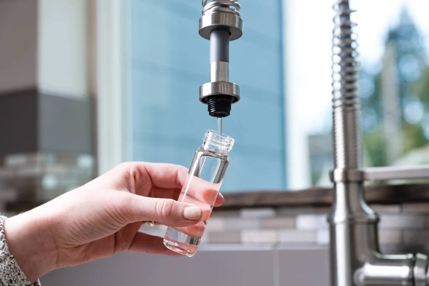 Hand holding glass test bottle with small amount of water, filling from a tall industrial kitchen faucet