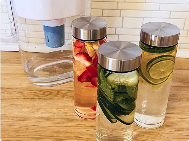 White Brita pitcher in background with Elite filter, and 3 reusable glass bottles with silver metal tops holding water with suspended lemon, cucumber and orange slices.