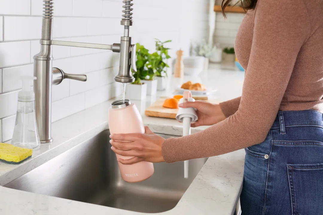 Woman filling off-white reusable water bottle from tall silver faucet.