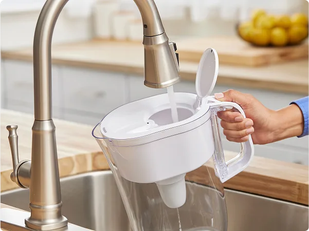 Hand holding white Brita pitcher while filling from a sink with a tall brushed bronze faucet.