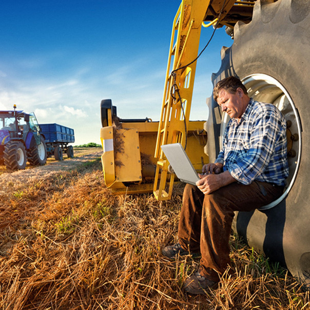 Ein Landwirt sitzt in einem großen Reifen und nutzt sein Laptop während der Arbeit auf dem Feld, im Hintergrund steht ein Traktor mit Anhänger