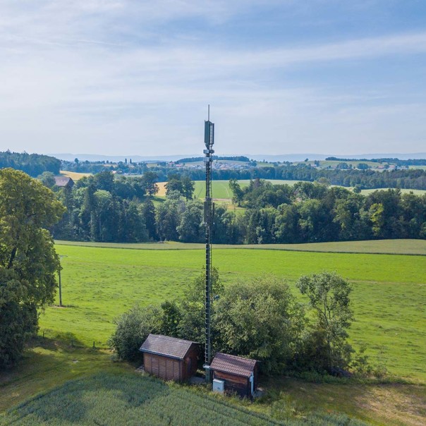 Mobilfunkmast neben zwei Häusern vor grüner Landschaft mit Bäumen. Der 5G-Betrieb gelingt ohne Umweltbelastungen.