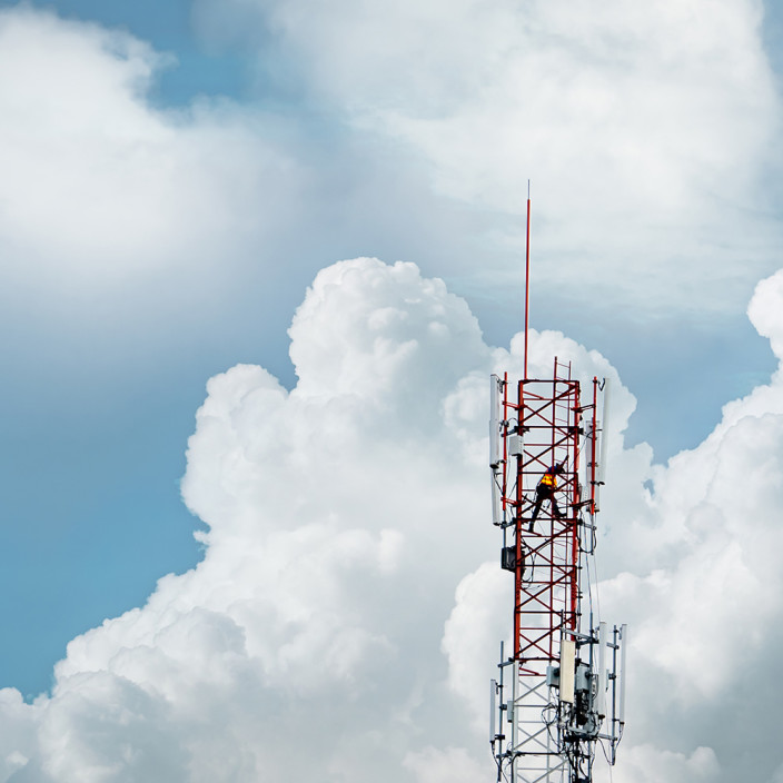 Mann steht auf einem Mobilfunkmast, der vor Wolken in den blauen Himmel ragt. Wir erklären, wie Politik, Behörden und Betreiber die neue 5G-Technik ausbauen.