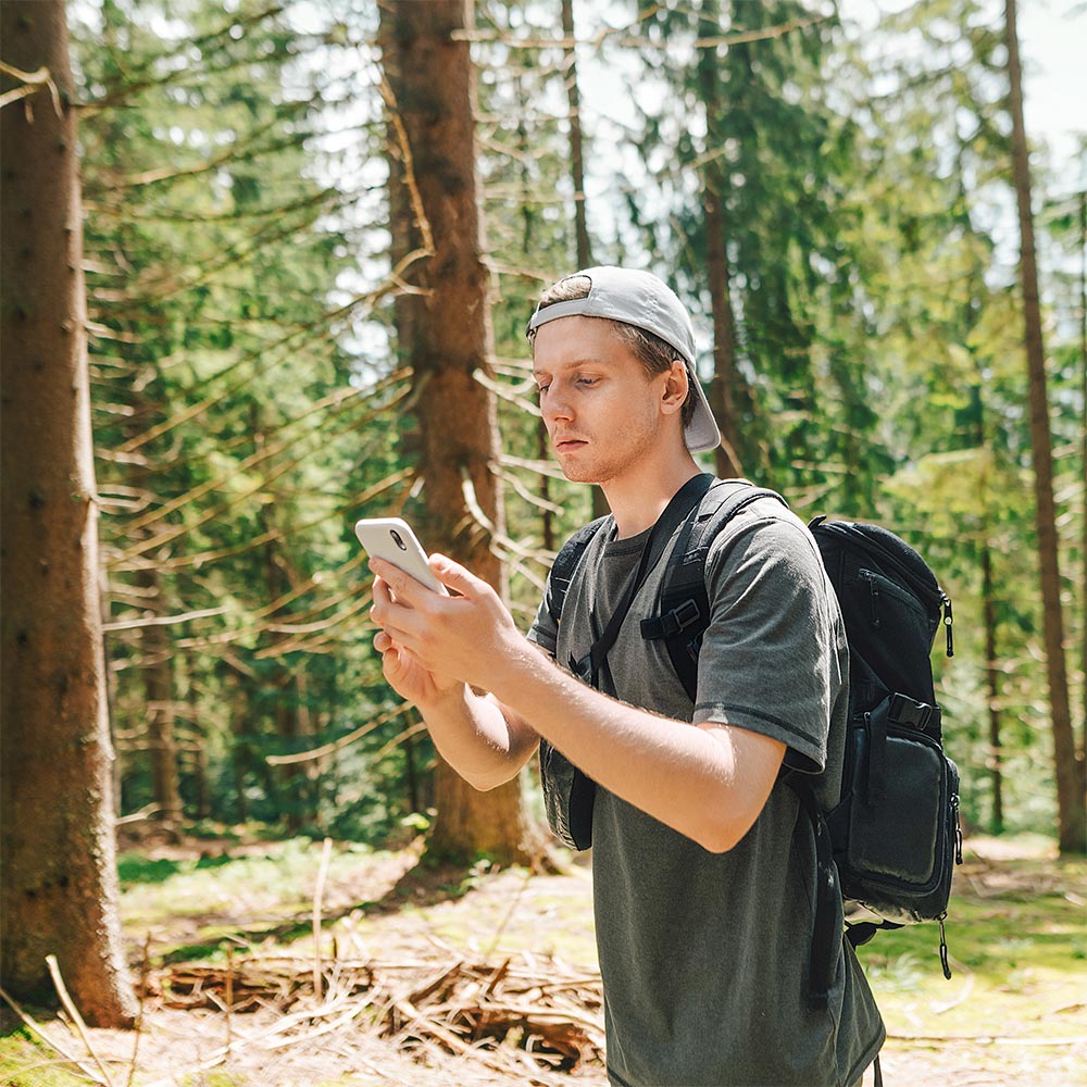 Mann mit Rucksack steht auf Waldlichtung und schaut auf sein Smartphone. Mitteilungen mit 5G schneller senden und empfangen – selbst mitten im Wald. 