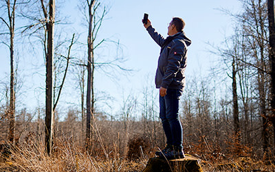 Mann steht im Wald auf einem Holzstumpf und misst mit einem Gerät in der Luft haltend Funk.