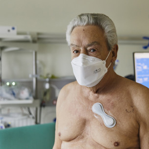 Älterer Mann ist in einem Krankenhaus-Zimmer mit Maske und hat ein Vitaldaten-Pflaster auf der Brust.