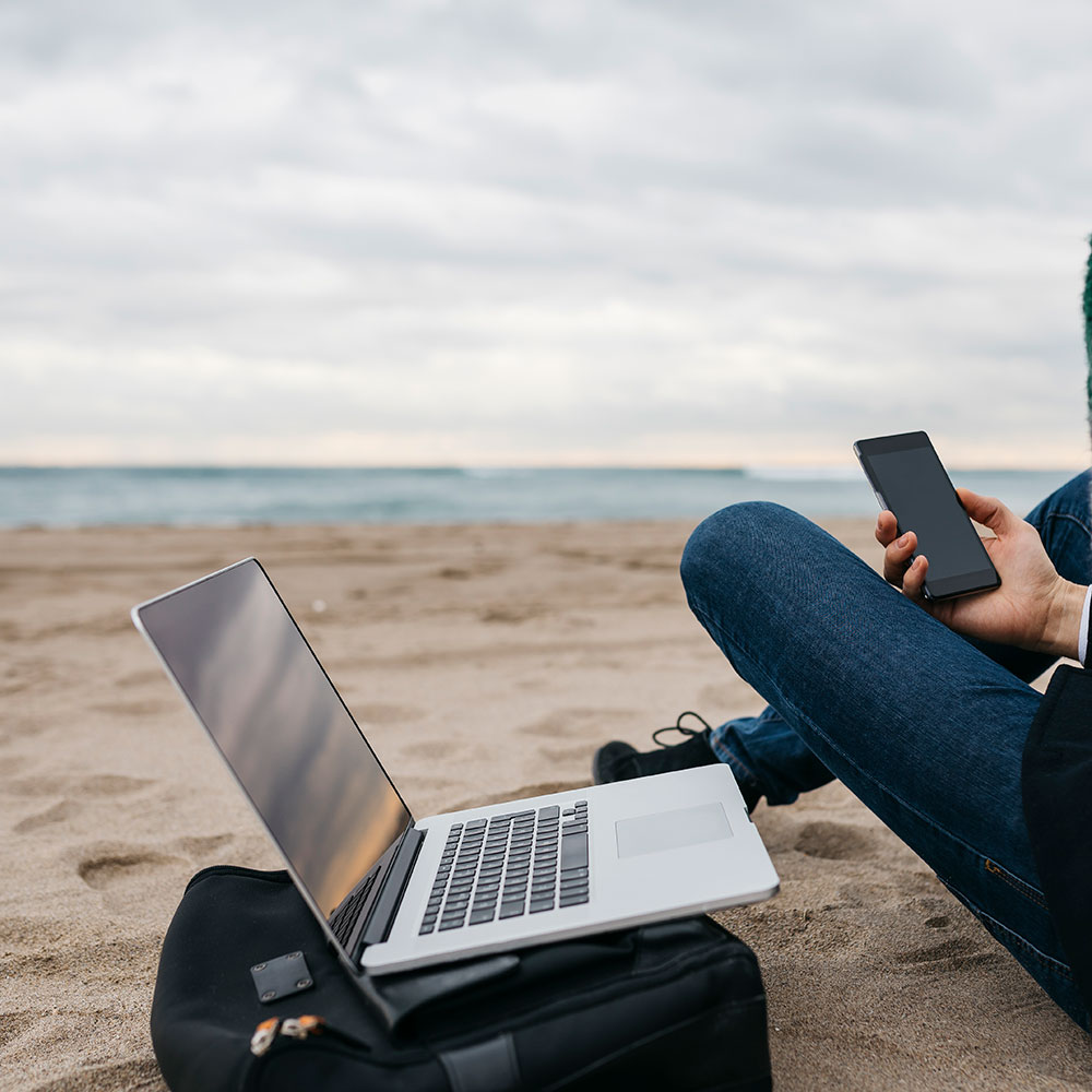 Person sitzt am Strand neben aufgeklapptem Laptop und hält Smartphone in der Hand. Strahlung gibt es in vielen Bereichen des Alltags. 