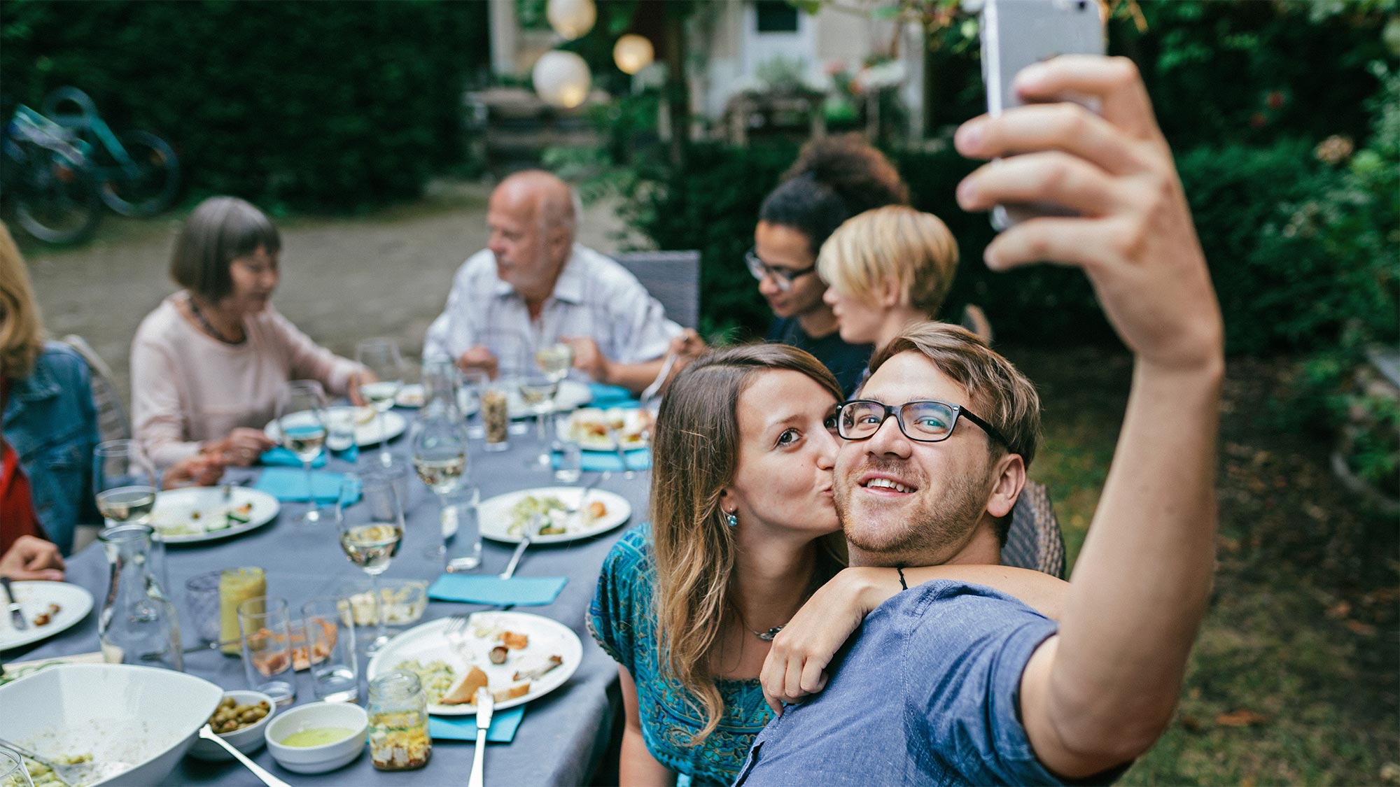 A man is sitting at a festive outdoor table taking a selfie while a woman is kissing him on the cheek. Private moments and messages remain private even with and thanks to 5G. 