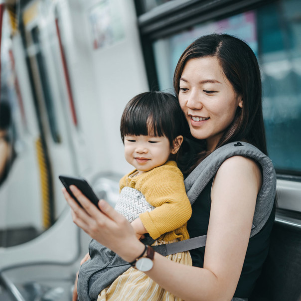 A woman is carrying a baby in a carrier bag on the subway, both looking at her cell phone. All forms of mobility will benefit from the new 5G technology. 