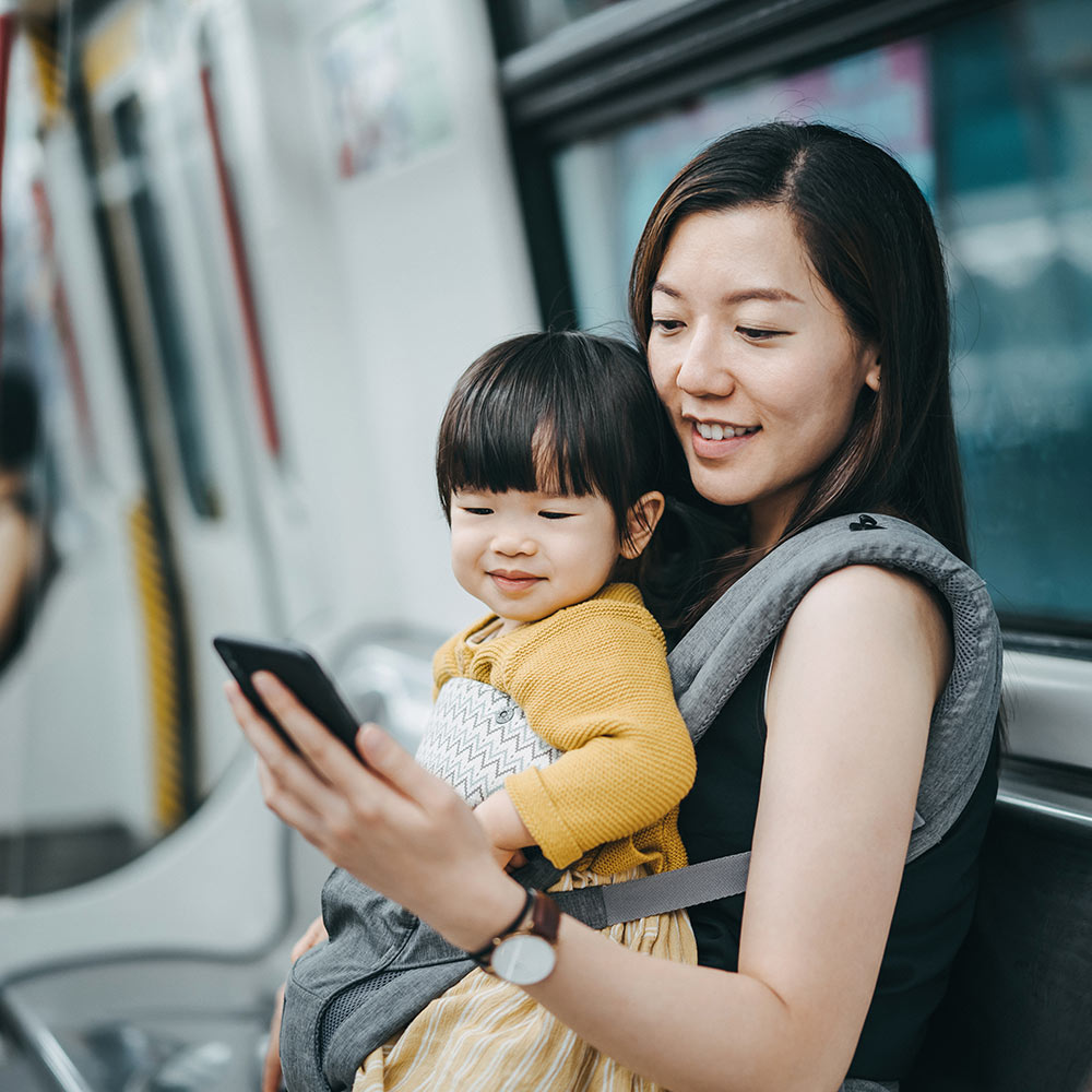 Frau trägt Baby in Tragetasche in der U-Bahn, beide schauen auf ihr Smartphone. Von der neuen 5G-Technologie werden alle Mobilitätsformen profitieren. 