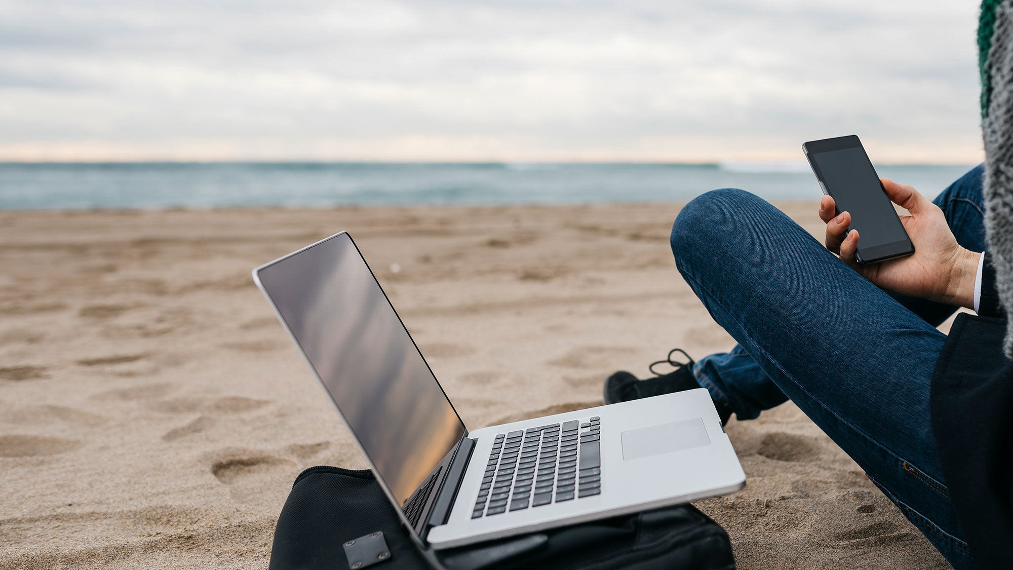 Person sitzt am Strand neben aufgeklapptem Laptop und hält ein Smartphone in der Hand. Strahlung gibt es in vielen Bereichen des Alltags. 