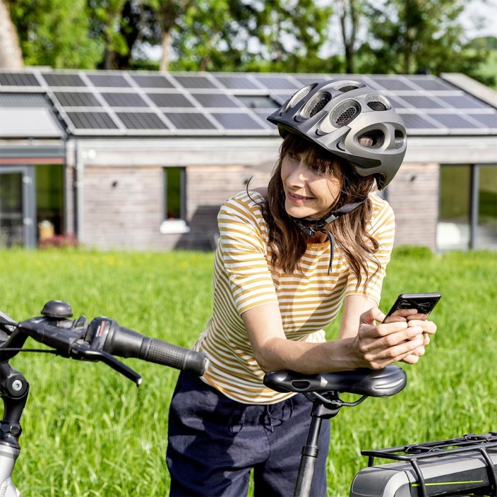 A cyclist is standing in front of a green meadow and is holding her mobile device in her hands. The 5G operation succeeds without environmental impact.