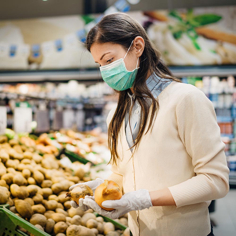 A woman in a supermarket is wearing a mouth and nose protection and gloves while standing in front of vegetable shelf. The fear that 5G spreads the corona virus is a myth. 