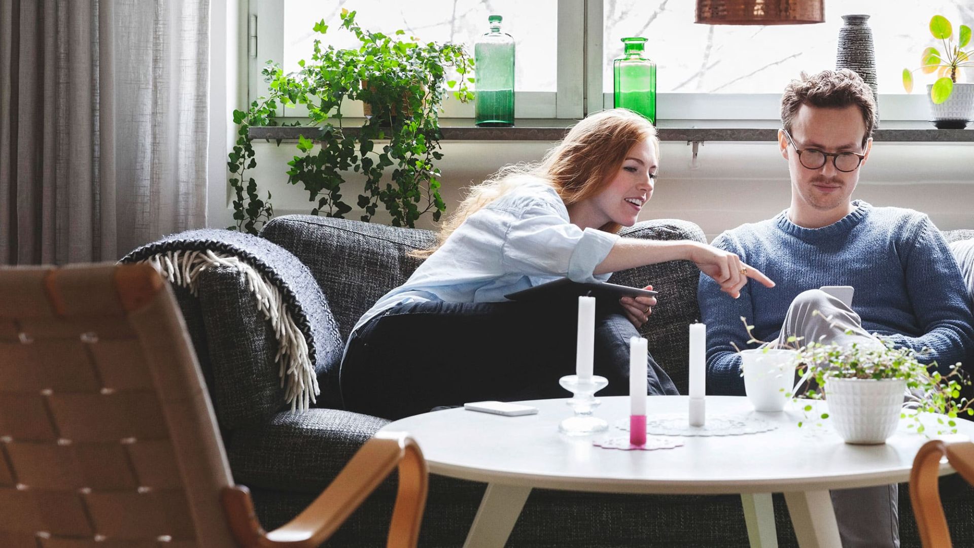 Eine Frau und ein Mann sitzen auf dem Sofa im Wohnzimmer. Der Mann hält ein Handy in der Hand. Die Frau zeigt auf das Display.