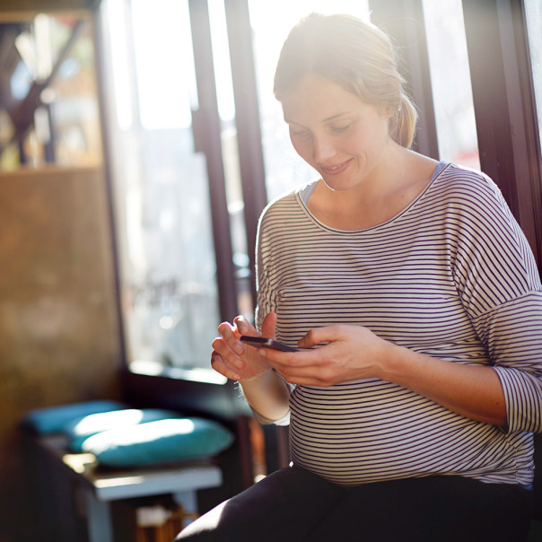 Schwangere Frau schaut lächelnd auf ihr Smartphone. Bisherige Studien konnten keine negative Wirkung von Mobilfunk auf ungeborene Babys oder die Fruchtbarkeit feststellen. 