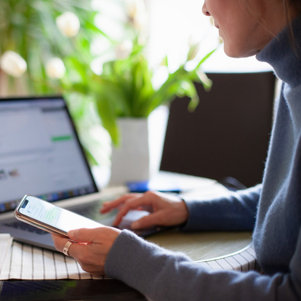 Frau sitzt an Arbeitsplatz, hält ein Smartphone und bedient währenddessen einen Laptop. Schneller Datenaustausch und nahtlose Kommunikation sind entscheidend für die Wettbewerbsfähigkeit der deutschen Wirtschaft. 