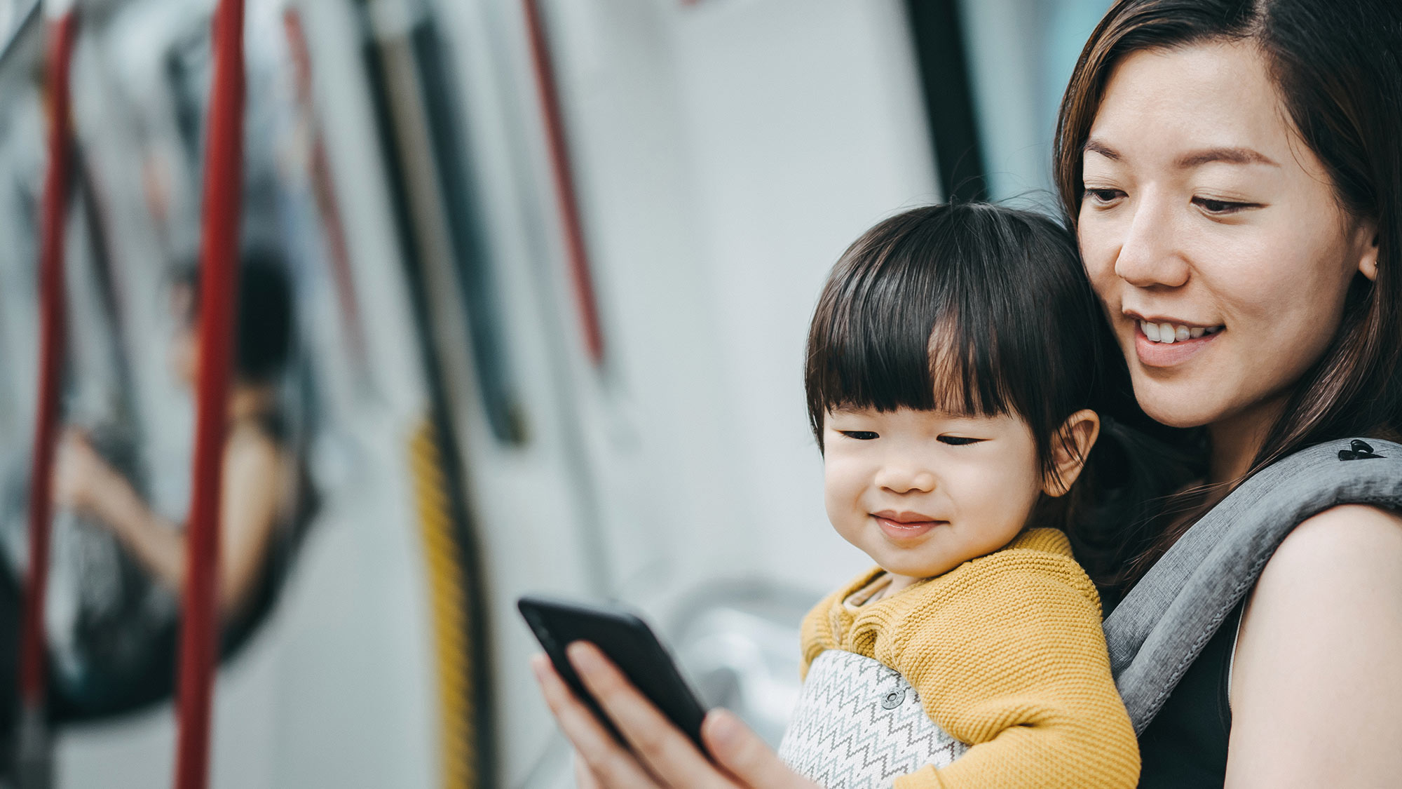 A woman is carrying a baby in a carrier bag on the subway, both looking at her cell phone. All forms of mobility will benefit from the new 5G technology. 