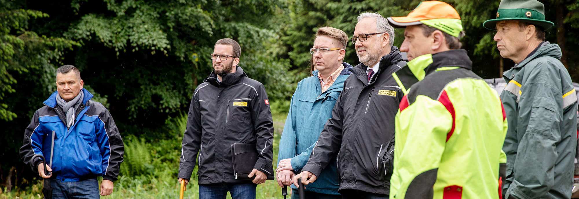 Herren mit Regenjacken und Schirmen stehen im Wald