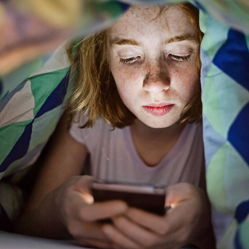 Mädchen mit Sommersprossen schaut auf ihr Smartphone unter der Bettdecke. Studien finden keine Belege für Schlaflosigkeit und Krankheiten durch elektromagnetische Felder. 