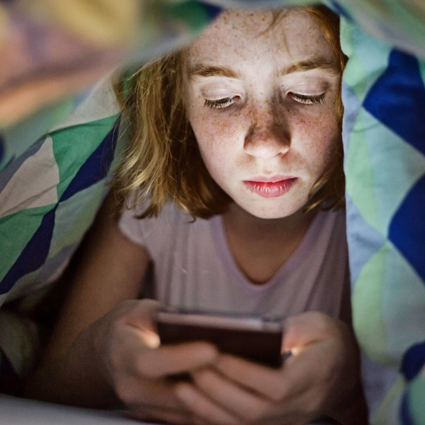 A girl with freckles is looking at a mobile device under the covers of her bed. Studies find no evidence of insomnia and diseases caused by electromagnetic fields. 