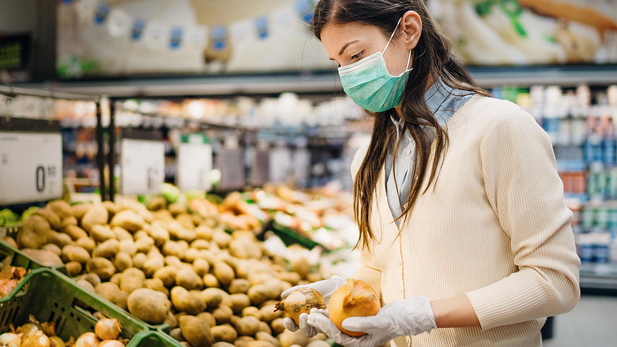 Frau im Supermarkt trägt einen Mund-Nasen-Schutz und Handschuhe während sie vor Gemüseregal steht. Die Angst, 5G verbreite das Coronavirus, ist ein Mythos. 