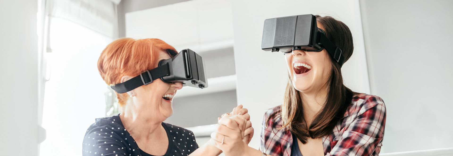 An older and a younger woman are sitting on the sofa at home wearing virtual reality glasses and are laughing.