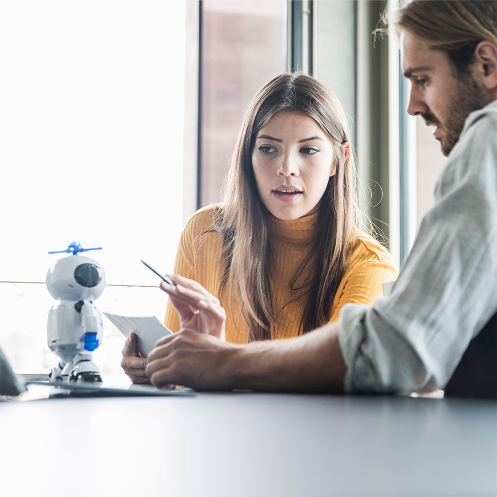 Frau und Mann sitzen an Konferenztisch und richten Blick auf einen kleinen Roboter. 5G und Künstliche Intelligenz stärken sich gegenseitig. Durch das Zusammenspiel werden die Technologien unseren Alltag bereichern und erleichtern.