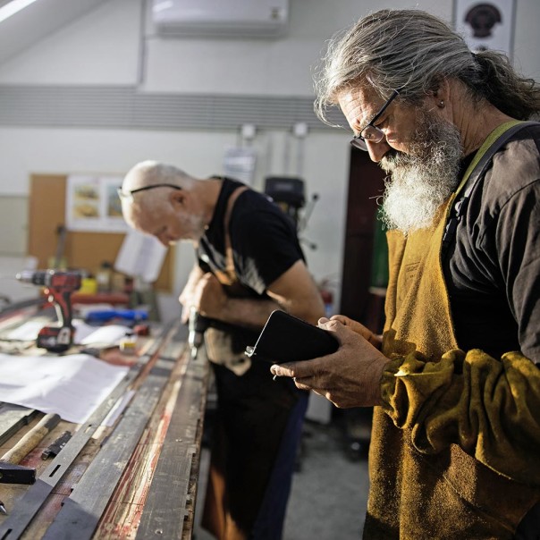 A man is standing in front of a workbench and is operating his mobile device while his colleague in the background is holding a drill in his hands. Digitalization will change the labor market. 