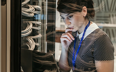 Frau in Serverraum schaut prüfend auf Laptop.